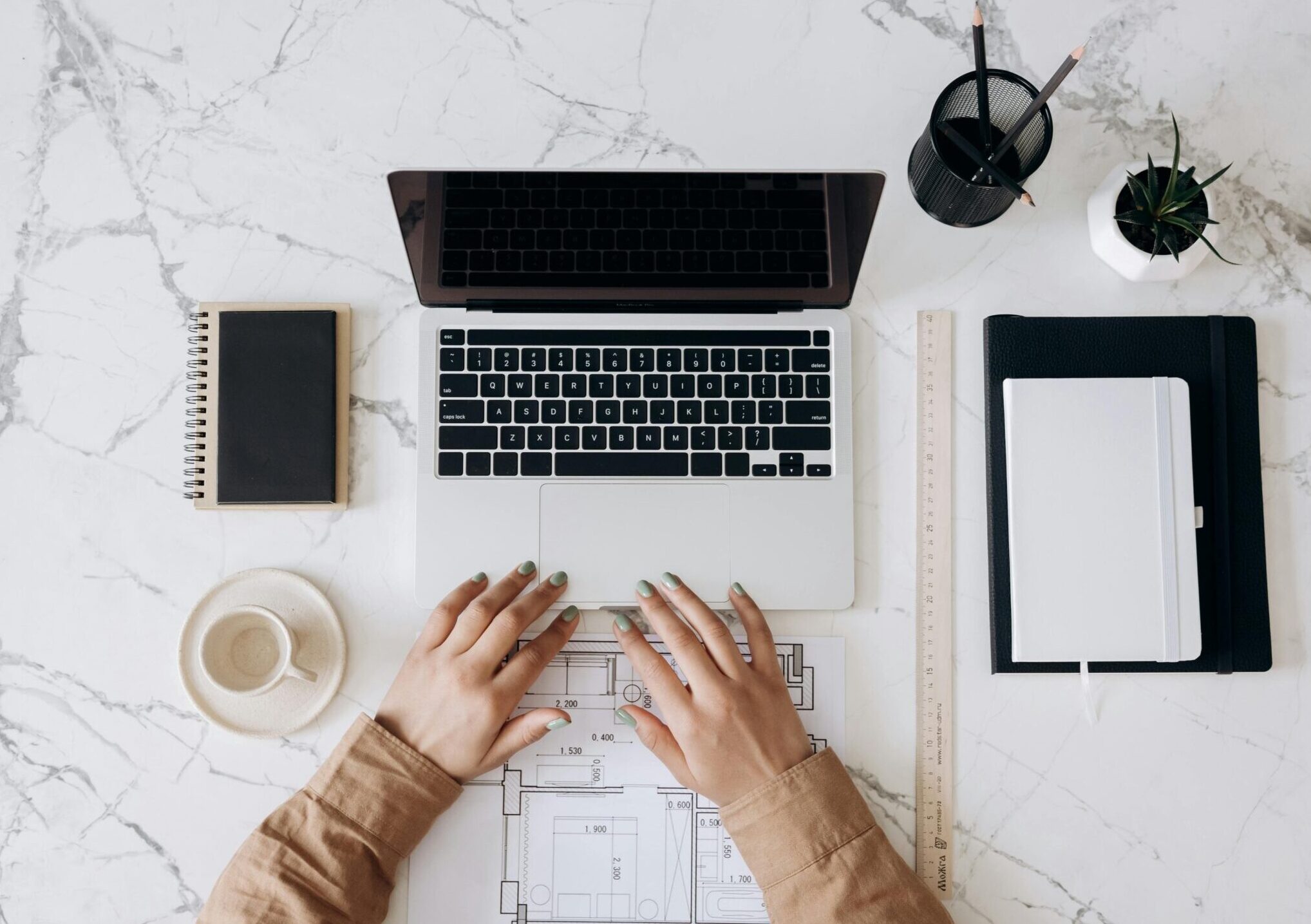 Top view of a stylish home office desk with a laptop, planner, and coffee cup, showing hands on a blueprint.