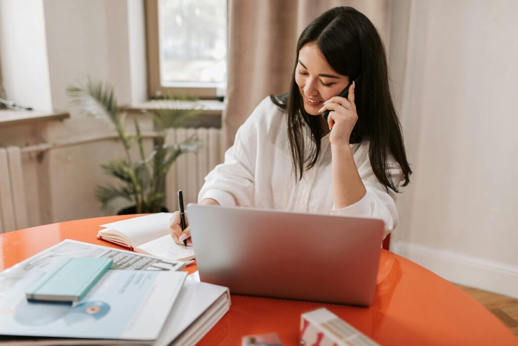 Young woman multitasking with laptop and phone in a modern home office setting.